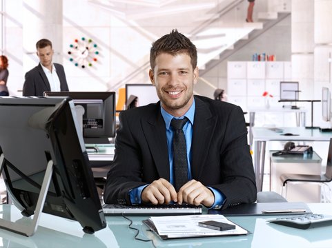 Happy young rookie caucasian businessman at business office with computer, sitting behind desk, smiling, looking at camera, suit and tie.