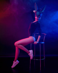 a beautiful girl in a bodysuit poses in a photo Studio on a dark background in neon light. On the face of a rabbit mask