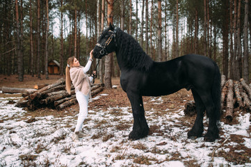 Magnetic young female in grey fur coat and white trousers stands with a big black horse outside