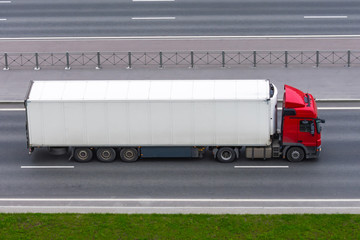 Truck with a white container on a trailer rides on a city highway, aerial side view.