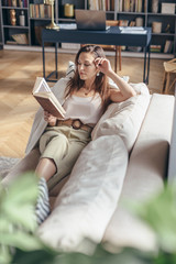 Young woman at home lying on couch and reading book.