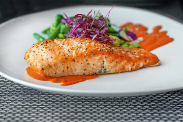 Closeup perfectly executed Norwegian salmon fillet steak with crispy fish skin, broccoli, young beans and spicy red sauce on table in restaurant. Plate with a dish of Norwegian salmon fish.