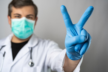 Caucasian man posing as a medical doctor in white overall or scrub with stethoscope around a neck and face mask. Showing peace sign with a hand in blue protective glove.