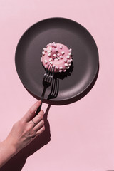 Light pink delicious donat with white marshmallows at the dark grey ceramic plate on the rosy background. Hand of woman with ring with freckles is going to eat donat by silver coloured fork. Vertical