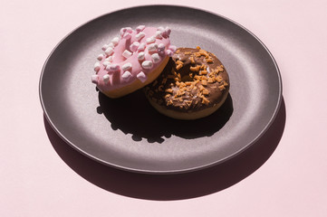 Two donats, one decorated with white marshmallows and another chocolate one with caramel-flavored curls on the the dark grey ceramic plate on pink background. Art minimalistic creative food concept