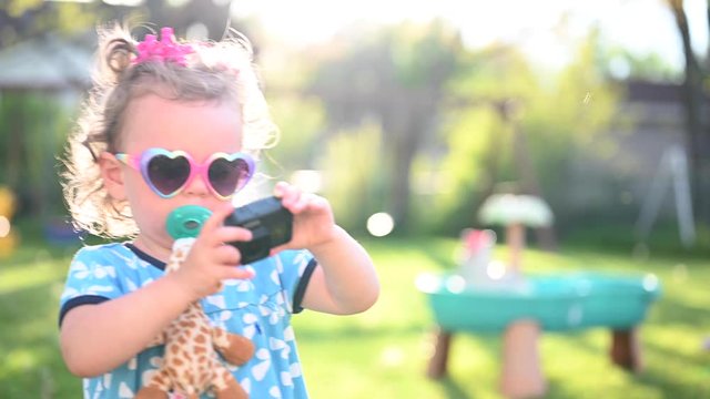 stylish little girl with pacifier and heart shaped sunglasses taking photos with action camera in backyard