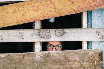Locked up guilty girl in glasses emotional portrait. Woman slavery. Werid strange unusual person looking through slit in boarded up window in old wooden abandoned ghost house. Female eyes expression.
