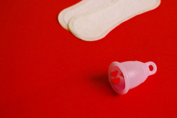 pink environmental silicone menstrual Cup in the foreground and two white pads in the back on a red background. choosing between a Cup and pads during menstruation