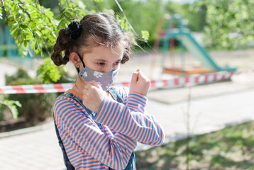 Cute little girl in a protective mask and stands with her arms crossed near a closed playground during coronavirus quarantine