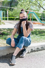 Teenage girl in protective mask sits on kerb near children's playground