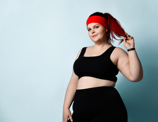 Concentrated plus-size girl in black sportswear stands looking aside thoughtfully, touching her...
