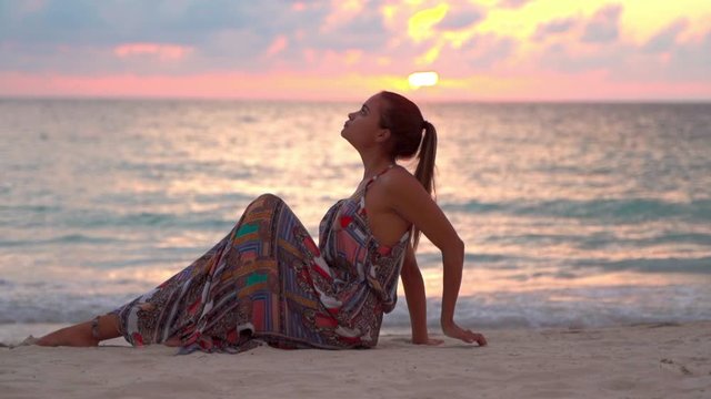 Slow motion of young woman relaxing at beach during sunset, beautiful female tourist wearing dress sitting on sand against sky - Montego Bay, Jamaica