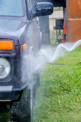 Car wash in the afternoon outdoors with a high-pressure apparatus. A powerful spray with water splashes away the dirt from the purple body of the car, from glass, wheels and tires.