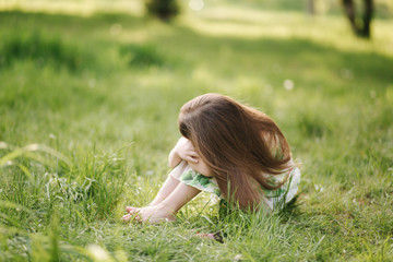Portrait of adorable little girl sits barefoot on the grass in the park. Happy kid on the fresh air