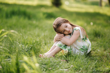 Portrait of adorable little girl sits barefoot on the grass in the park. Happy kid on the fresh air