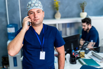 Male doctor talking over the phone. Doctor in blue scrubs. Sitting on table angle. Medical concept.