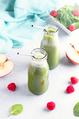 Two glass bottles of green smoothie with reusable drinking straws with spinach, raspberry and apple on concrete table, still life. Healthy freshly blended fruits and herbs smoothie, top view
