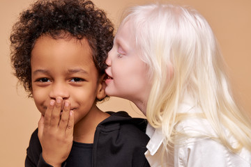 albino girl with pale skin and white hair color tells a secret in the ear of multiracial african boy. cute boy with black skin attentively listen to secret. isolated - obrazy, fototapety, plakaty