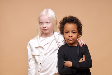 two friendly black afro american and albino children stand together isolated over beige background, diverse positive boy and girl stand hugging. children concept