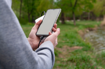 Man uses smartphone while charging from the power bank on the river's bank. Modern technology concept. Selective focus.