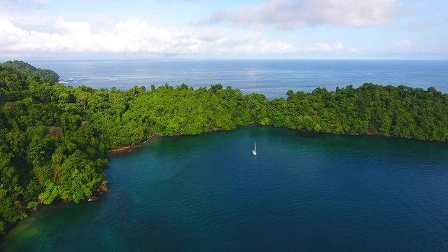 Decending Aerial View of Anchored Sailboat Near Brincanco Island (Coiba Park) in Blue Lagoons with Dense Jungle Trees and Rock Cliff Shore