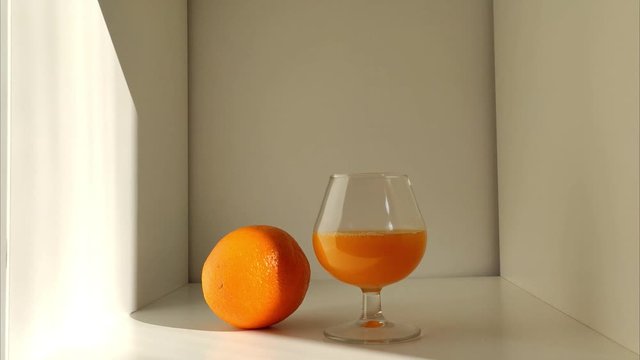 Timelapse de un zumo de naranja en una estanter&iacute;a blanca. El sol va girando por la escena. 