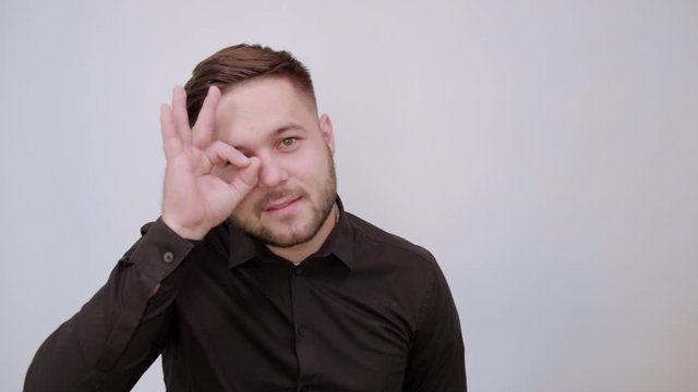 Young Bearded Dark Haired Man In Black Stylish Shirt On White Background, Happy Male Smiles, Made Ok Sign With Fingers, Holds It To His Eyes. All Right. The Concept Of Successful And Affluent People