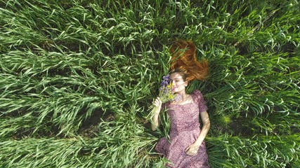 Beauutiful young red haired woman lying on green summer field in sunny weather. Aerial view of happy girl in purple dress relaxing and smelling flowers on sunny grass in park. Summer holidays concept.