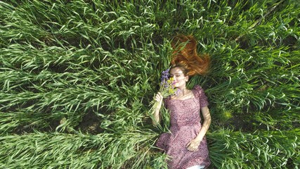 Beauutiful young red haired woman lying on green summer field in sunny weather. Aerial view of happy girl in purple dress relaxing and smelling flowers on sunny grass in park. Summer holidays concept.