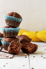 stack of homemade сhocolate muffins with banana in blue paper cups  on white, wooden background. Fresh bananas near it.