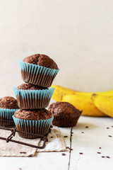stack of homemade сhocolate muffins with banana in blue paper cups  on white, wooden background. Fresh bananas near it.