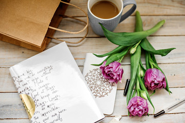 creative arrangement  beautiful dark pink tulips, a paper bag, a notebook, a pen  and a cup of coffee on a wooden background. Flat lay. Selective focus.