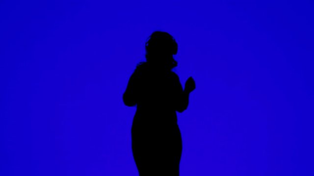 Silhouette of a slender woman wearing headphones and dancing to the music on a blue background