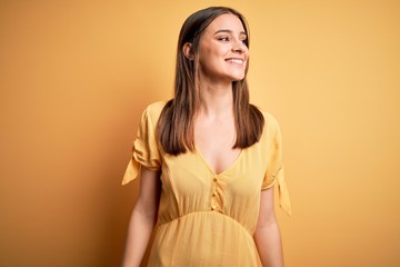 Obraz premium Young beautiful brunette woman wearing casual dress standing over yellow background looking away to side with smile on face, natural expression. Laughing confident.