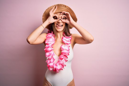 Young beautiful brunette woman on vacation wearing swimsuit and Hawaiian flowers lei doing ok gesture like binoculars sticking tongue out, eyes looking through fingers. Crazy expression.