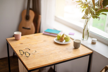 close-up photo of wooden table at home. home desk for freelance work. eyeglasses, cup of coffee on table. guitar in the background