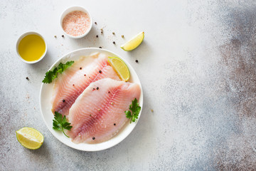 Raw tilapia fillet with lime and spices on a gray stone background - obrazy, fototapety, plakaty