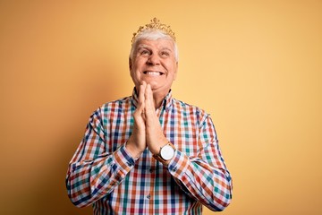 Senior handsome hoary man wearing golden crown of king over isolated yellow background begging and...