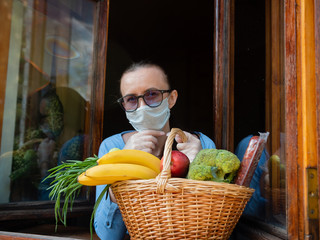 A sad woman in a protective mask, standing at the window, holds a basket of products from volunteer donations