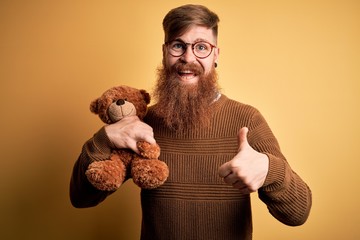 Redhead Irish man with beard hugging teddy bear stuffed animal over yellow isolated background...