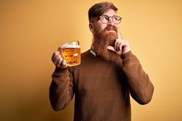 Irish redhead man with beard drinking a glass of refreshing beer over yellow background serious face thinking about question, very confused idea