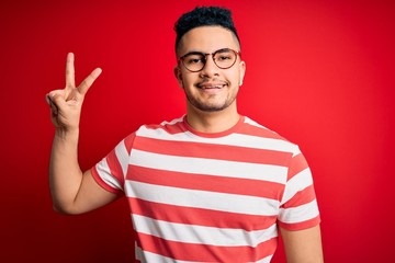 Naklejka premium Młody przystojny mężczyzna ubrany w casualową koszulkę w paski i okulary na na białym tle czerwonym tle uśmiechnięty, patrząc do kamery pokazując palce robi znak zwycięstwa. Numer dwa.