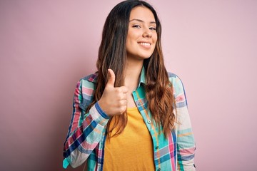 Young beautiful brunette woman wearing casual colorful shirt standing over pink background doing happy thumbs up gesture with hand. Approving expression looking at the camera showing success.