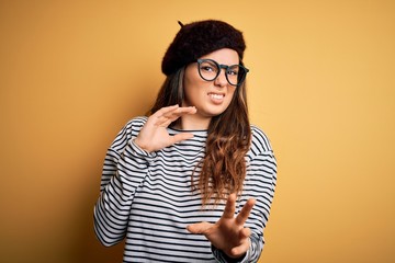 Young beautiful brunette woman wearing french beret and glasses over yellow background disgusted...