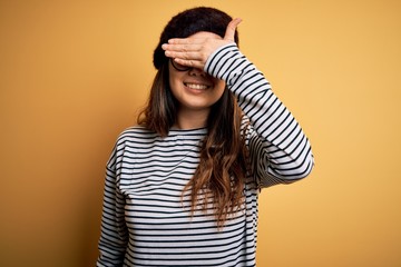 Young beautiful brunette woman wearing french beret and glasses over yellow background smiling and laughing with hand on face covering eyes for surprise. Blind concept.