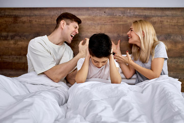 little child tired of quarrels mom and dad, kid sit between parents on bed closing his ears from screams