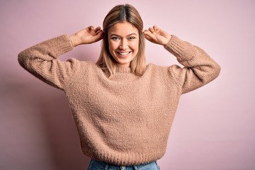 Young beautiful blonde woman wearing winter wool sweater over pink isolated background Smiling pulling ears with fingers, funny gesture. Audition problem