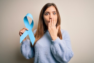 Young blonde woman holding prostate cancer awareness campaing blue ribbon cover mouth with hand...