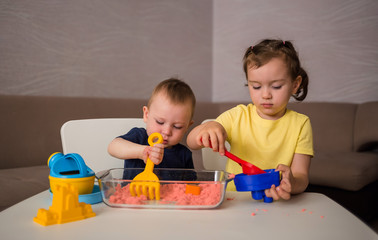 A boy and a girl play kinetic sand at a table in a room. A little girl pours into the mold in the...