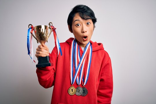 Young beautiful asian girl winner holding trophy wearing medals over white background scared in shock with a surprise face, afraid and excited with fear expression